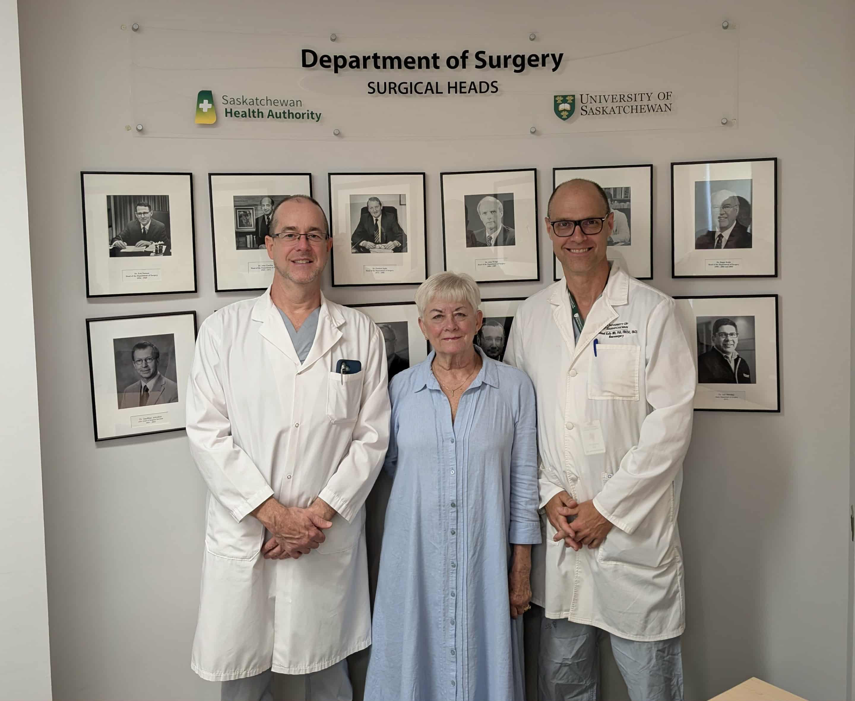 (L to R) Dr. John Shaw (MD), Nancy Keith, and Dr. Michael Kelly (MD) standing in front of the wall of Surgical Heads. (Photo by Jared Fingler)
